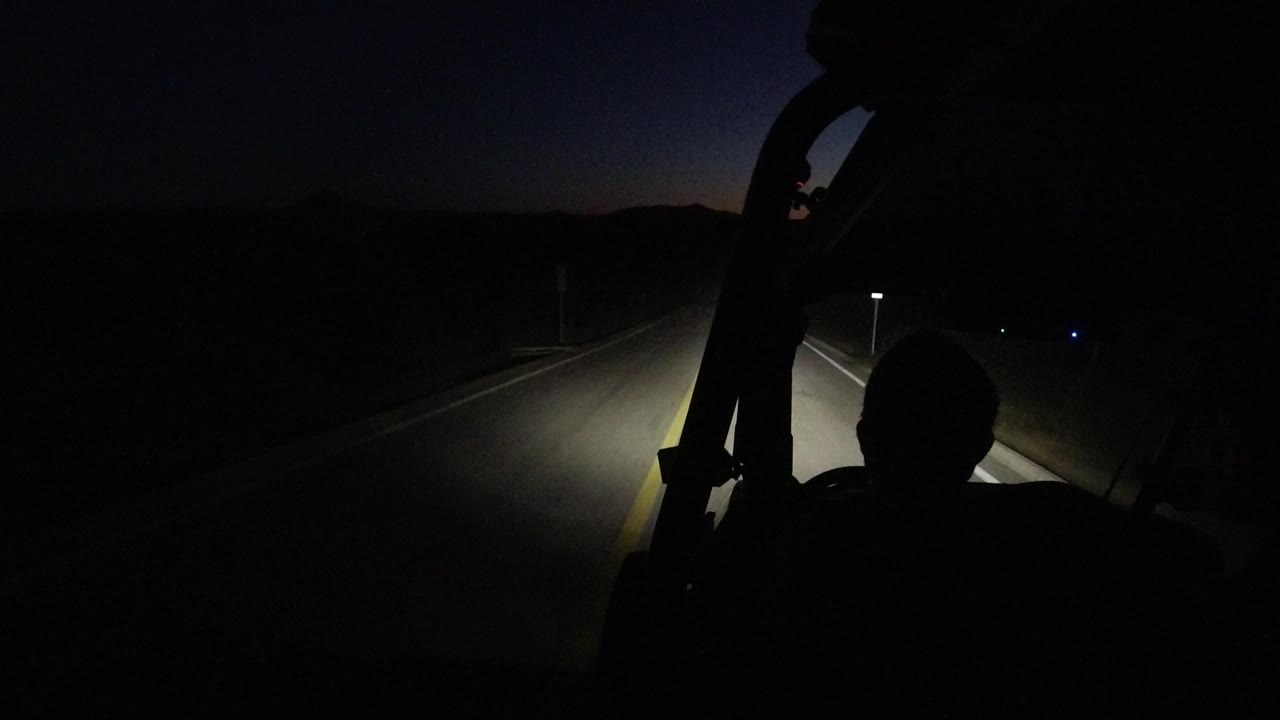 A dim Los Cabos roadway stretches ahead while the foreground cabin silhouette frames the headlight beam, with no recognizable people visible.