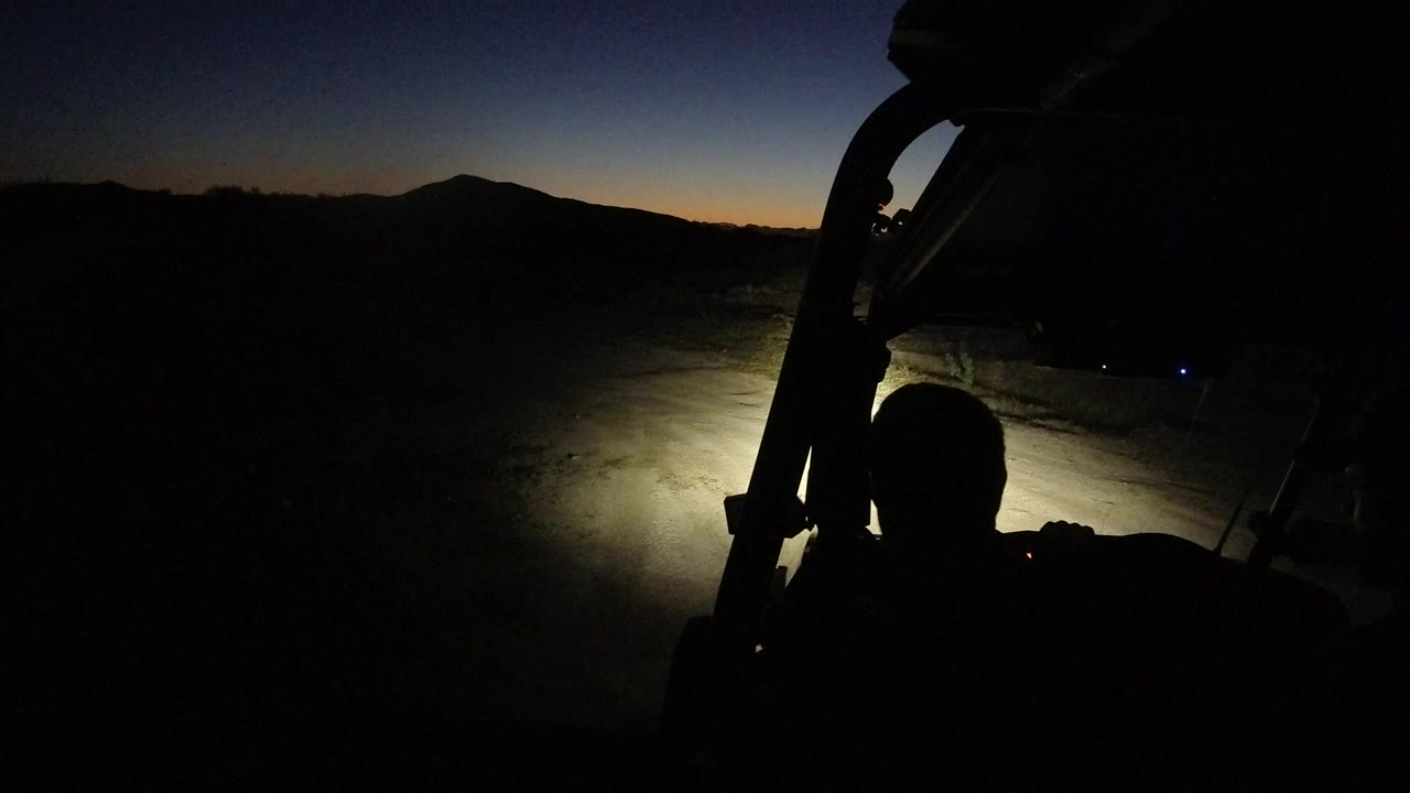 Opening frame in Los Cabos, Mexico showing a dark vehicle frame, headlight beam on desert ground, a mountain silhouette, and a silhouetted seated person.