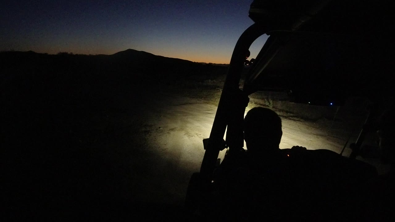 Los Cabos night desert frame with the headlight pool centered lower in view, dark foreground vehicle structure, a faint blue-orange sky, and a silhouetted person in front.