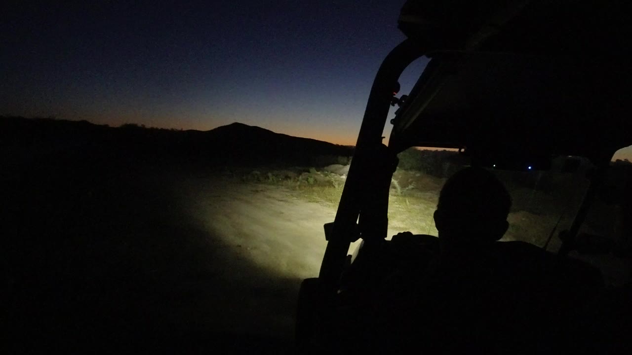 A silhouetted passenger and roll bar frame a headlit dirt road in Los Cabos, Mexico, with a low mountain horizon under a blue-black sky.