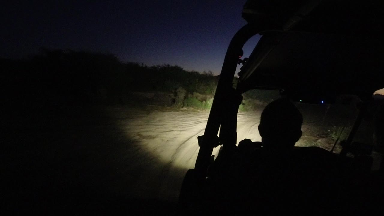 The Jeep cabin remains dark in Los Cabos, Mexico, with the driver's head in silhouette and the headlight beam spreading across the road edge and brush.