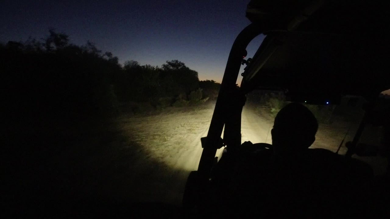 A later night-driving frame in Los Cabos shows the headlight beam centered on the dusty track, with the seated figure still in silhouette and scrub lining the road.