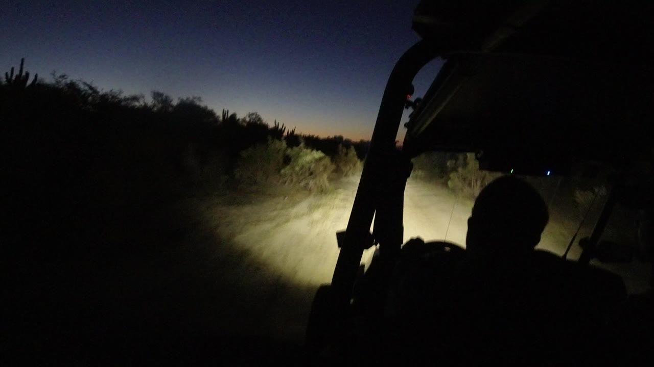 The cart continues along the Los Cabos dirt track, with the driver in shadow and the headlight cone falling across dust, roadside plants, and a narrow road edge.