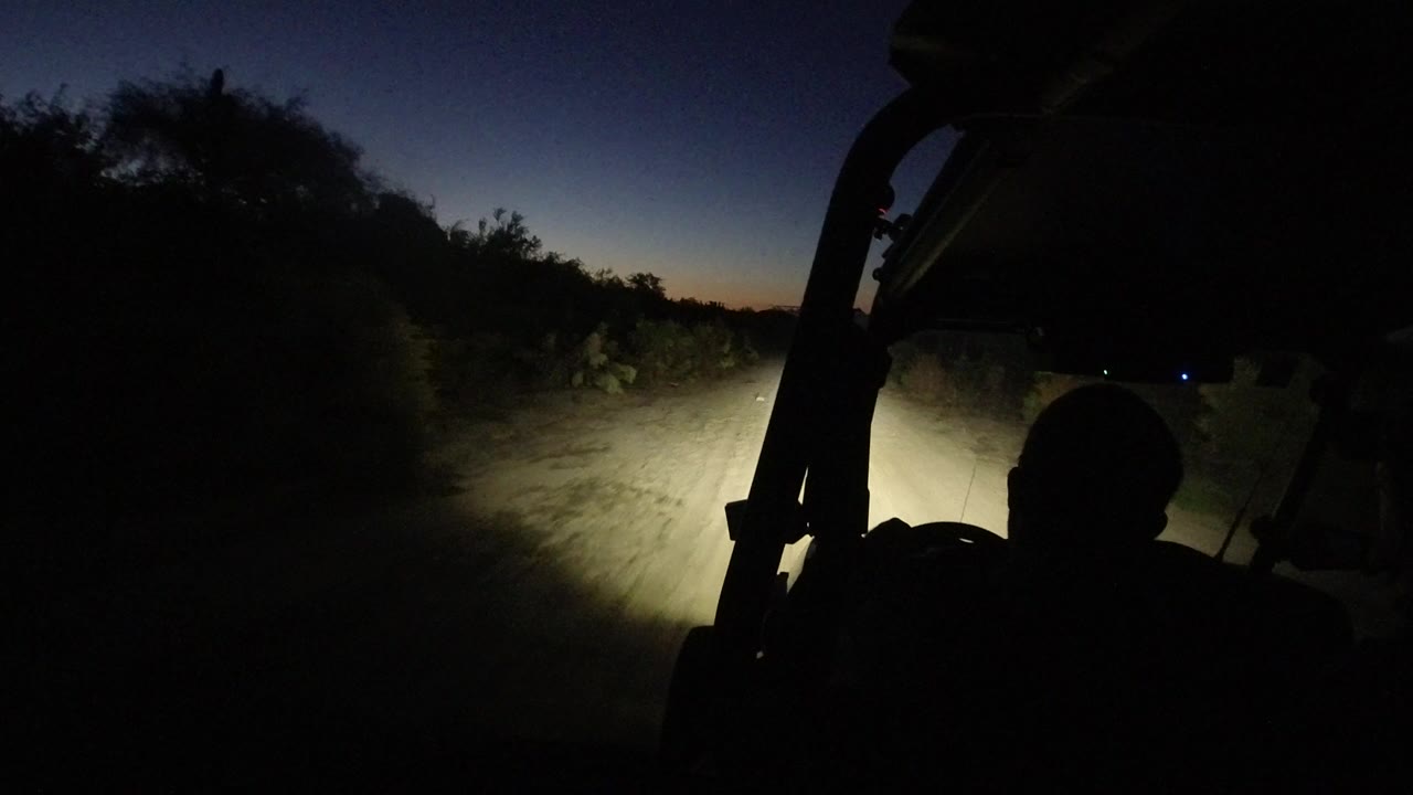 The Jeep continues along a dim dirt track in Los Cabos, Mexico, with the headlight beam centered on the road and a silhouetted vehicle frame at right.
