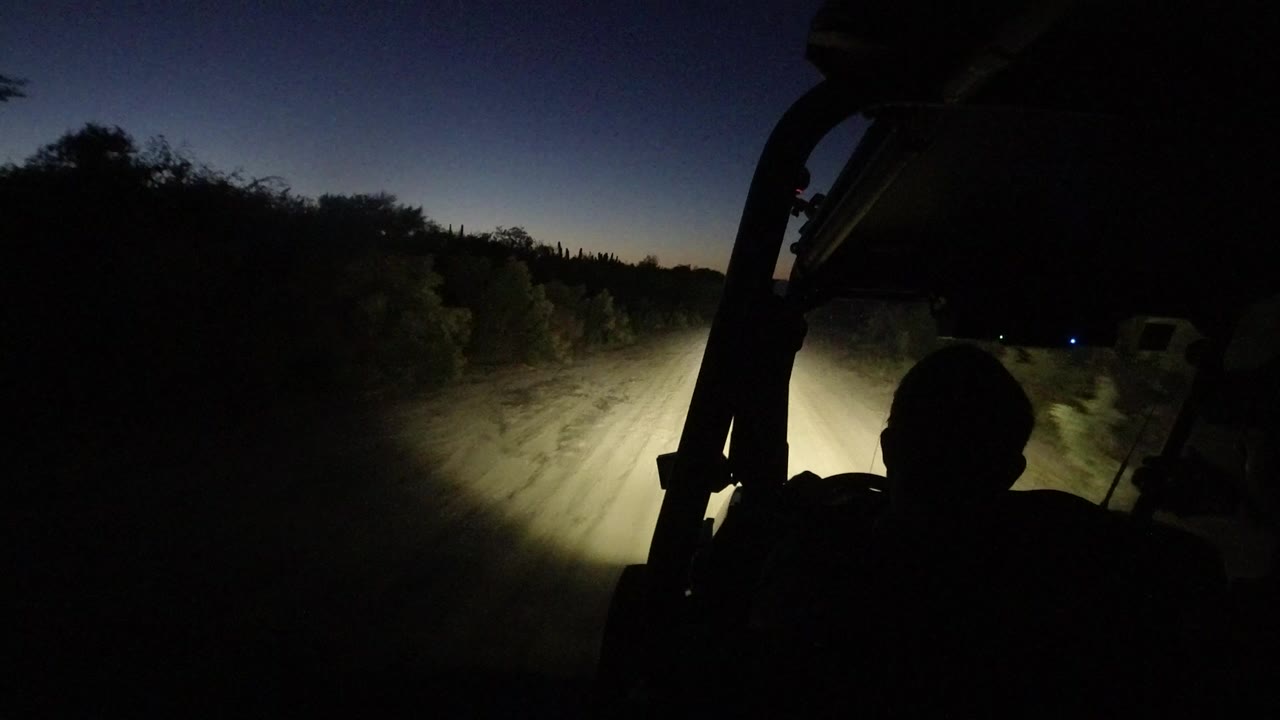 Dark Jeep interior facing a headlit dirt road at dusk.