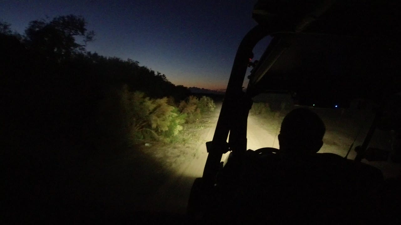 At 0:00 in Los Cabos, Mexico, a dark driver silhouette sits inside a Jeep as headlights light brush beside a narrow road under a fading blue sky.