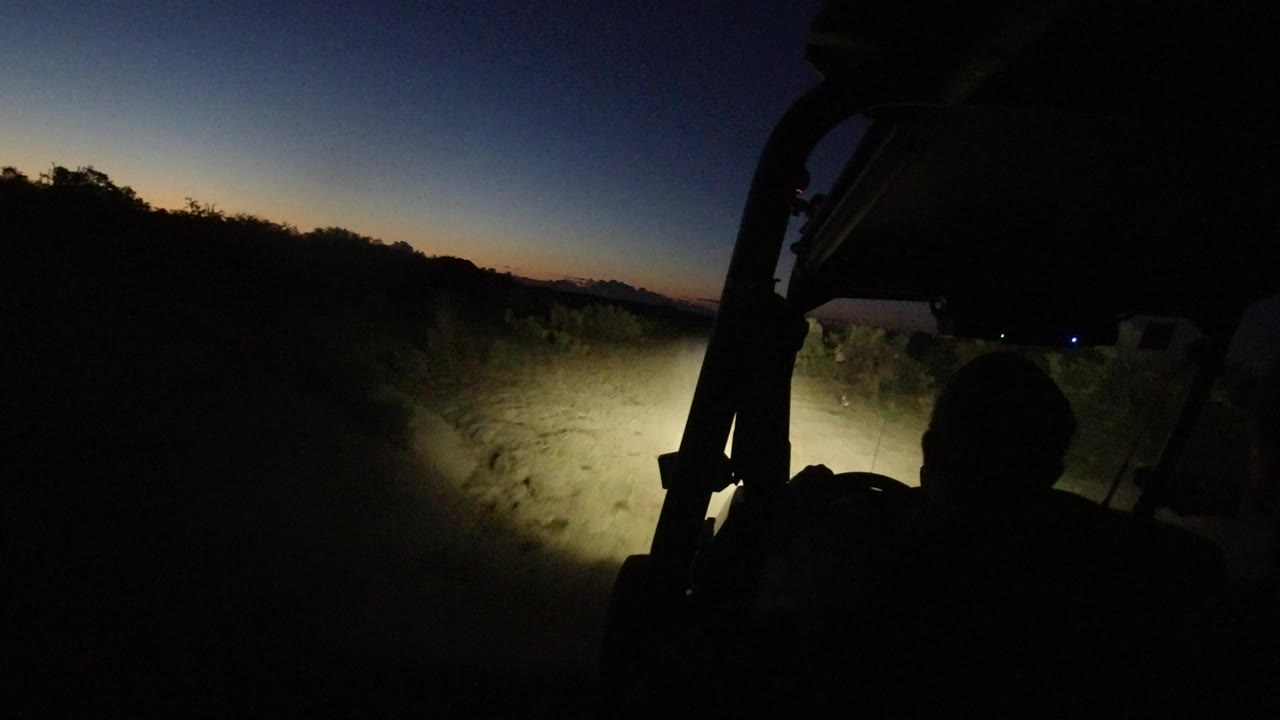 The second frame keeps the silhouetted passenger low in the foreground while the headlights pick out pale roadside plants under a blue dusk sky in Los Cabos, Mexico.