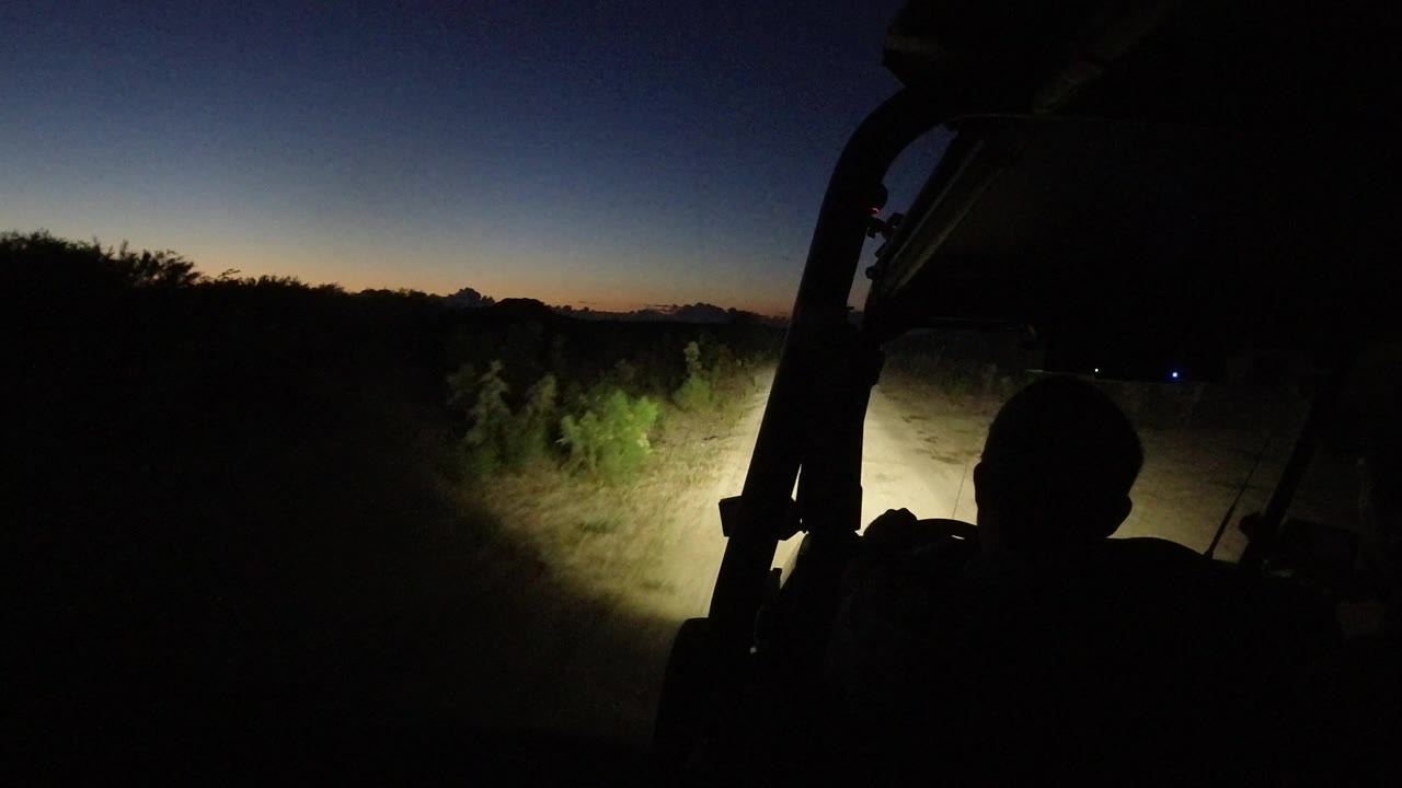The Los Cabos road remains mostly dark as the vehicle headlights catch pale brush on the right and the sky fades from orange at the horizon to deep blue above.