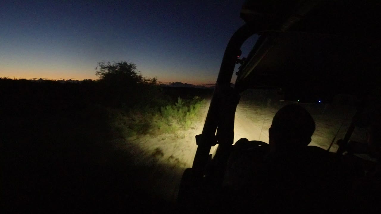 Inside or beside an off-road vehicle in Los Cabos, Mexico, headlights illuminate sandy brush under a dark blue and orange dawn sky with no identifiable faces visible.