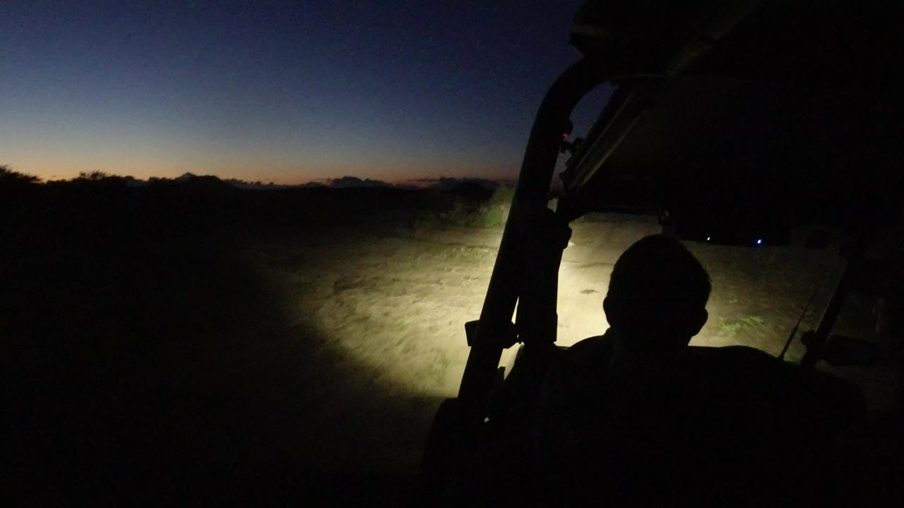 At 0:00 in Los Cabos, Mexico, a dark driver silhouette sits in the foreground while headlights illuminate a pale dirt track and low desert brush under a blue-black sky.
