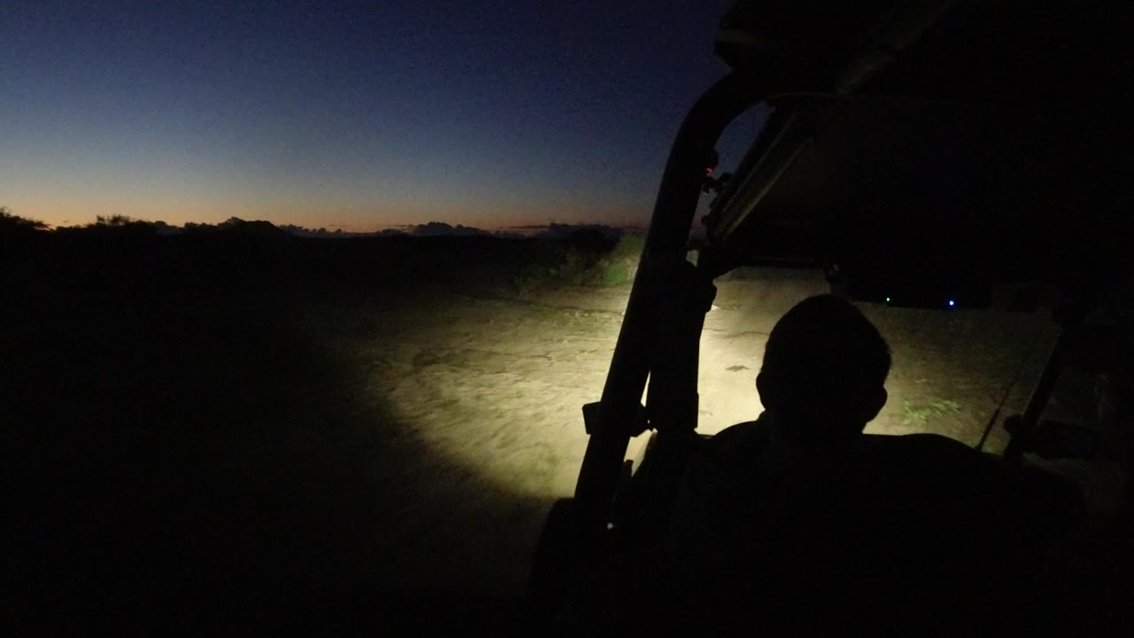At 0:01, the silhouetted driver remains centered against the vehicle frame as the headlights spread across dusty ground and low vegetation beneath a thin dusk horizon.