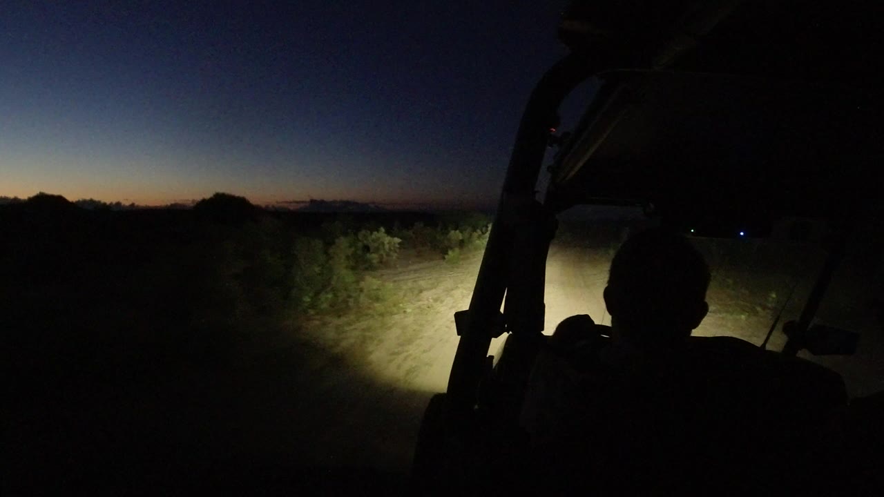 A silhouetted dune buggy interior faces a headlight-lit dirt road in Los Cabos, Mexico, with scrub brush and a blue-orange dawn horizon visible ahead.