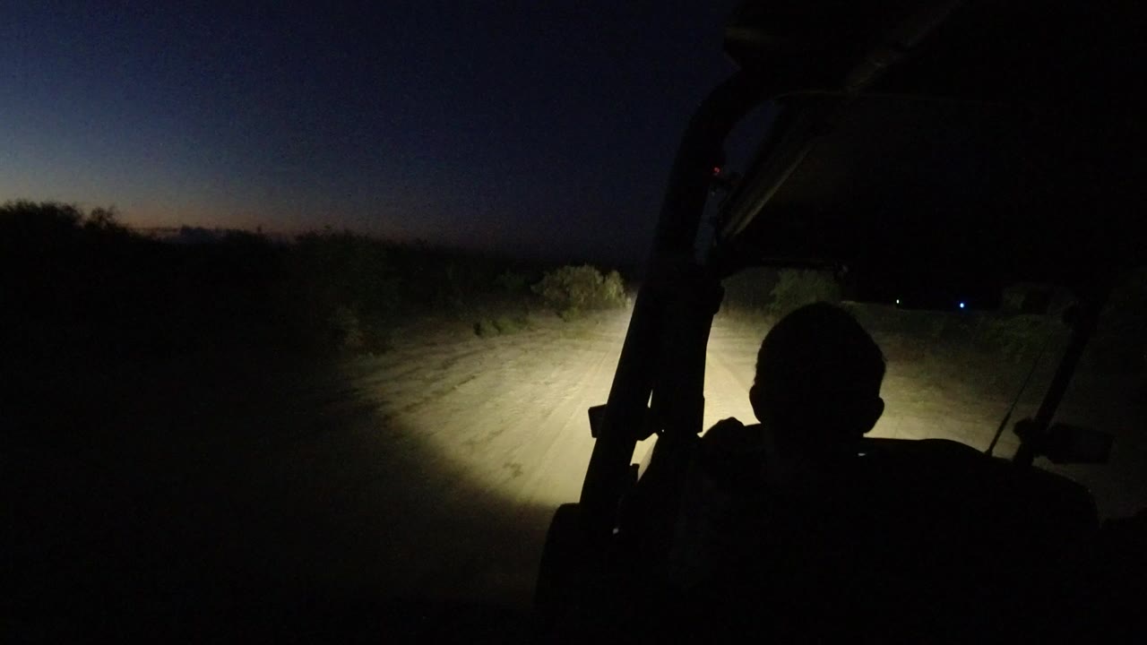 Headlights on a dark dirt road from behind a silhouetted vehicle occupant.