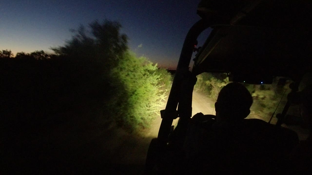 The vehicle view continues in Los Cabos, Mexico, with a dark human silhouette in the foreground and green brush glowing in the headlight beam beside the road.