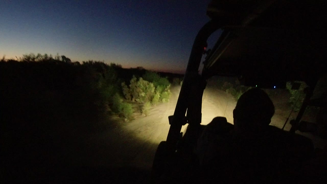A Los Cabos, Mexico dirt road is lit by vehicle headlights from an interior view, with dark brush on both sides, a faint horizon, and no recognizable faces visible.