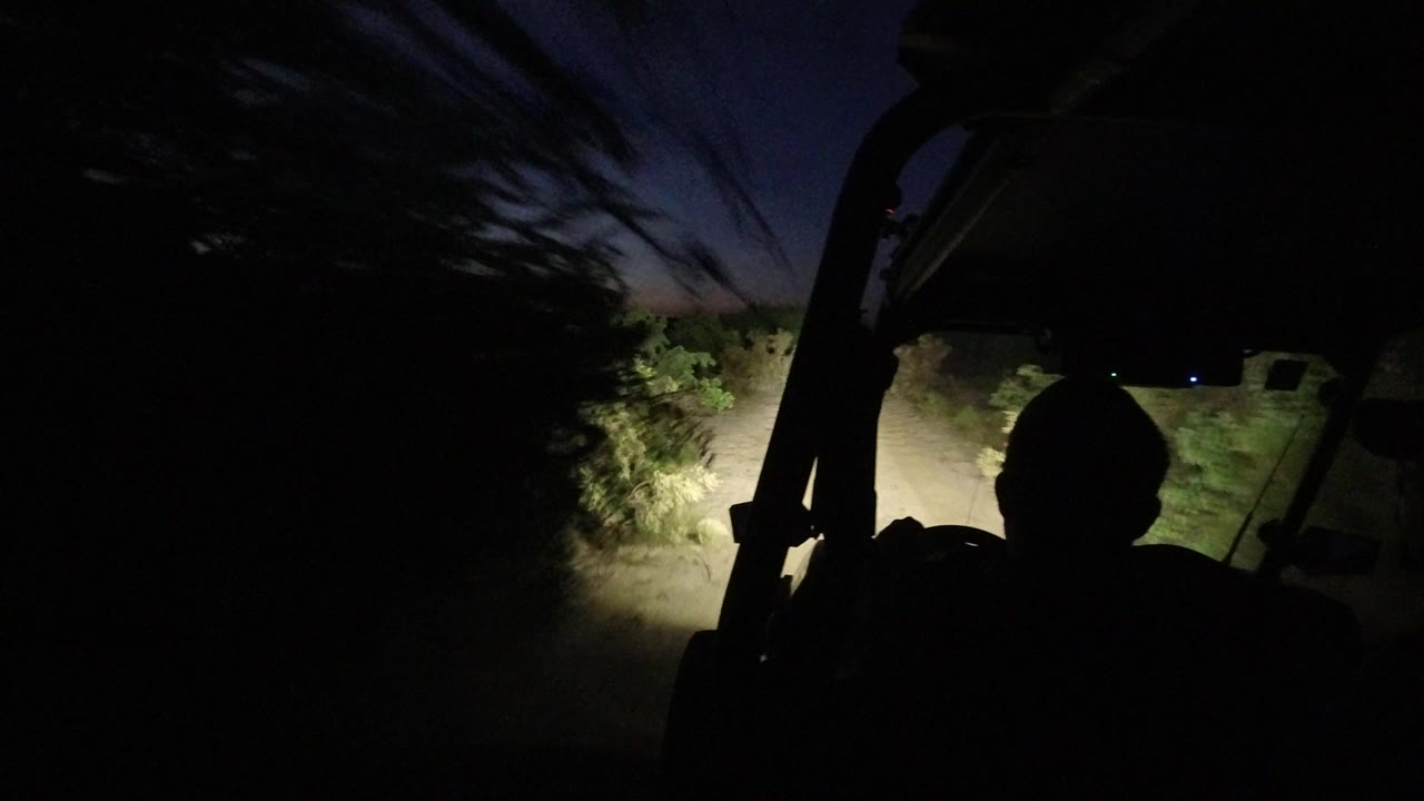 The driver remains in dark silhouette while the headlights catch roadside brush and dust under a fading blue sky in Los Cabos, Mexico.