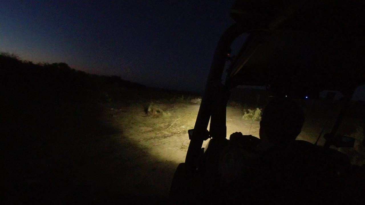At 0:00 in Los Cabos, Mexico, a dark forward vehicle view shows headlights illuminating uneven dirt and low brush under a nearly black sky, with only a non-recognizable silhouette inside the vehicle.