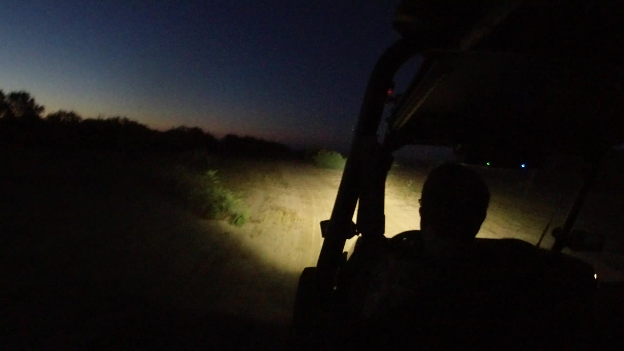A silhouetted driver and Jeep roll bar frame a headlight-lit dirt road in Los Cabos, Mexico, with low bushes and a fading blue sky ahead.