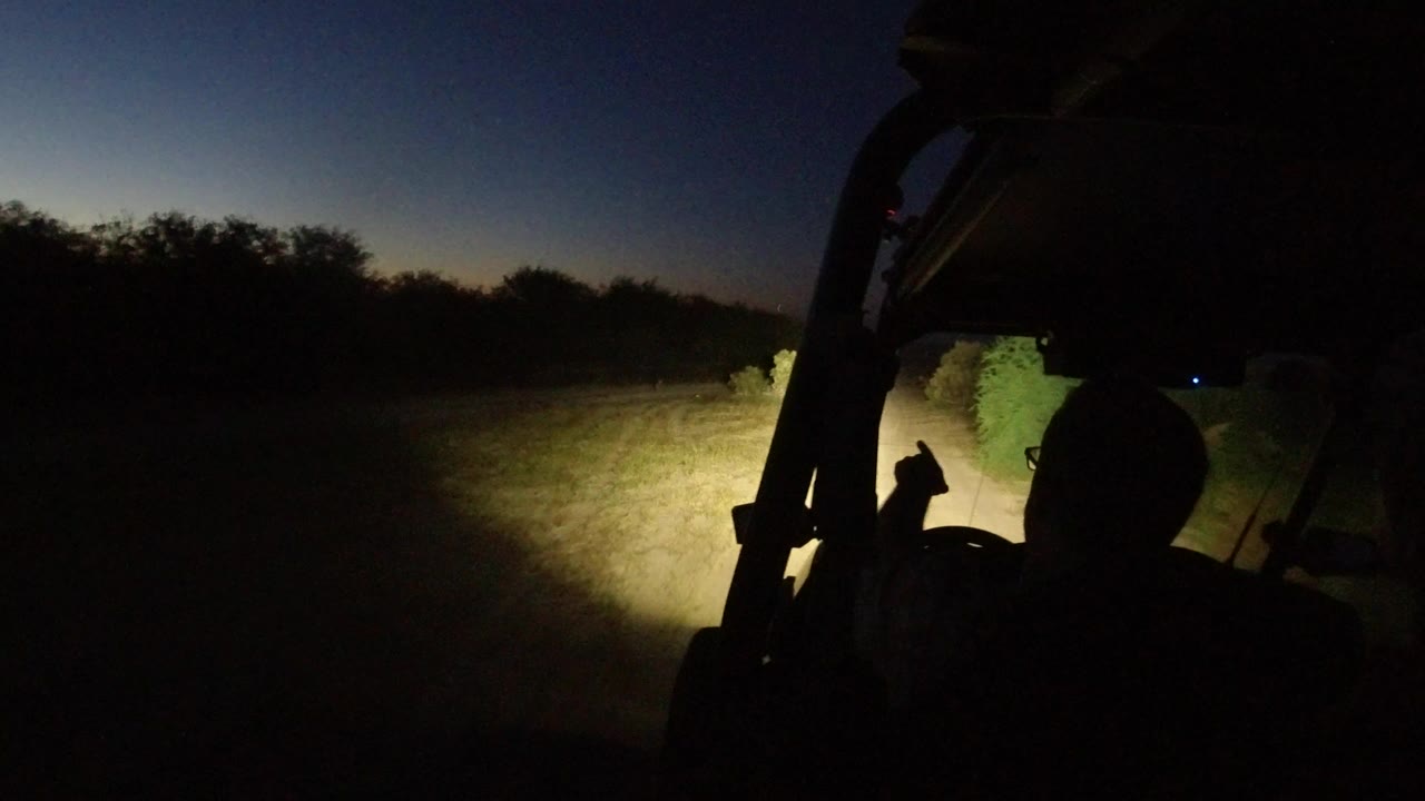 In Los Cabos, Mexico, a dark driver silhouette sits inside an open vehicle while headlights illuminate a dirt road and roadside brush under a fading blue sky.