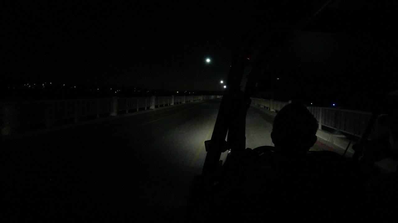 A low nighttime view from inside or near a Wrangler shows the Los Cabos bridge lane continuing forward, with white railings, small distant lights, and a dark foreground silhouette.