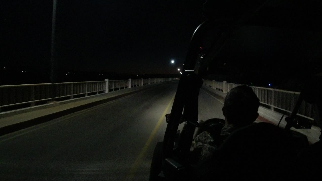 Los Cabos night driving frame with the road centered, pale bridge railings on both sides, a bright light ahead, and a person in silhouette at right foreground.