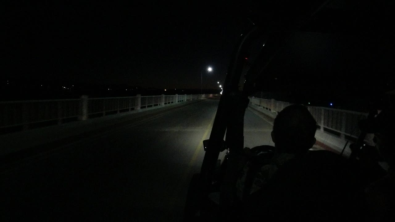 At 0:01 in Los Cabos, Mexico, the road remains mostly empty as the headlights reach toward a brighter streetlight and the foreground silhouette stays dark.