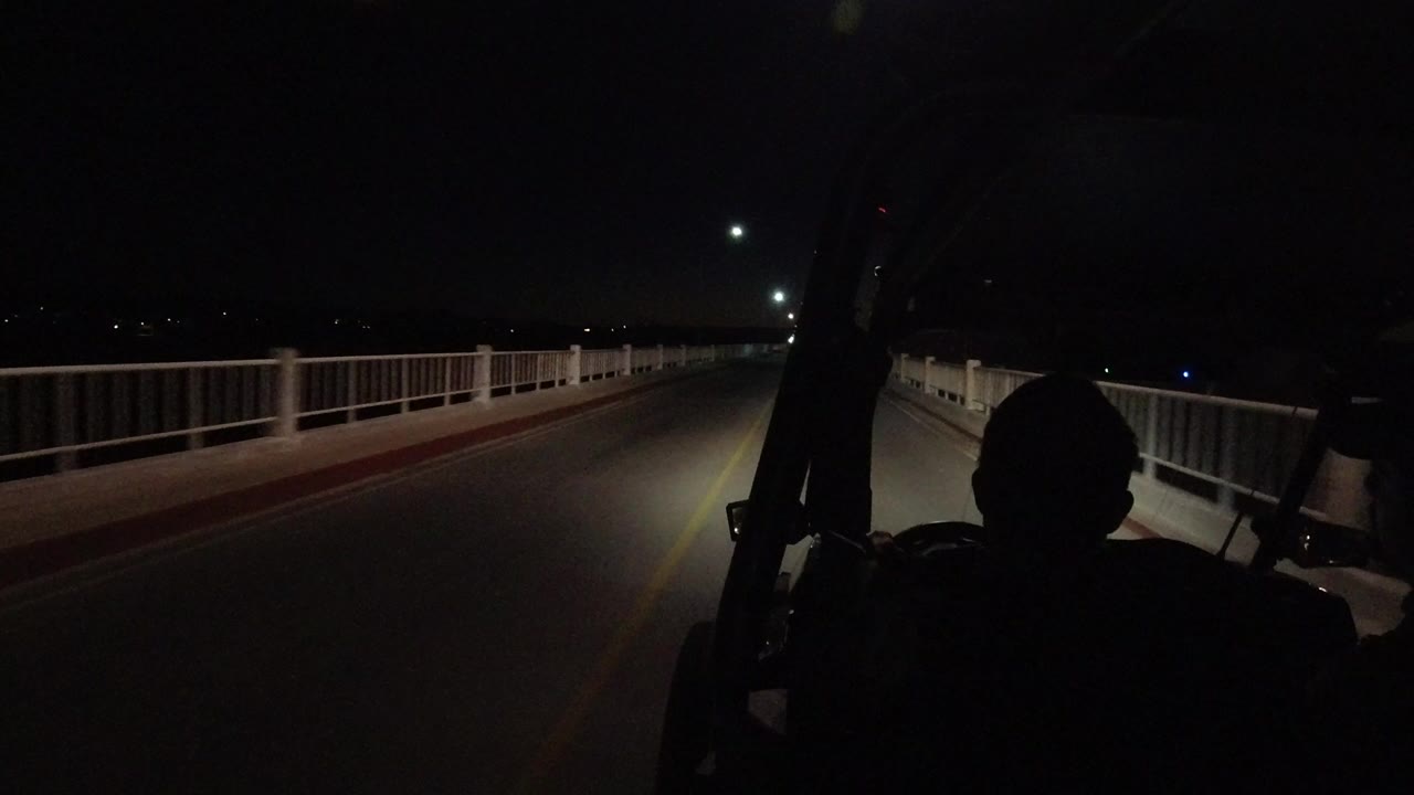 Start of a Los Cabos night drive showing a two-lane road, white bridge railings, headlight spill, streetlights, and a dark foreground silhouette inside the vehicle.