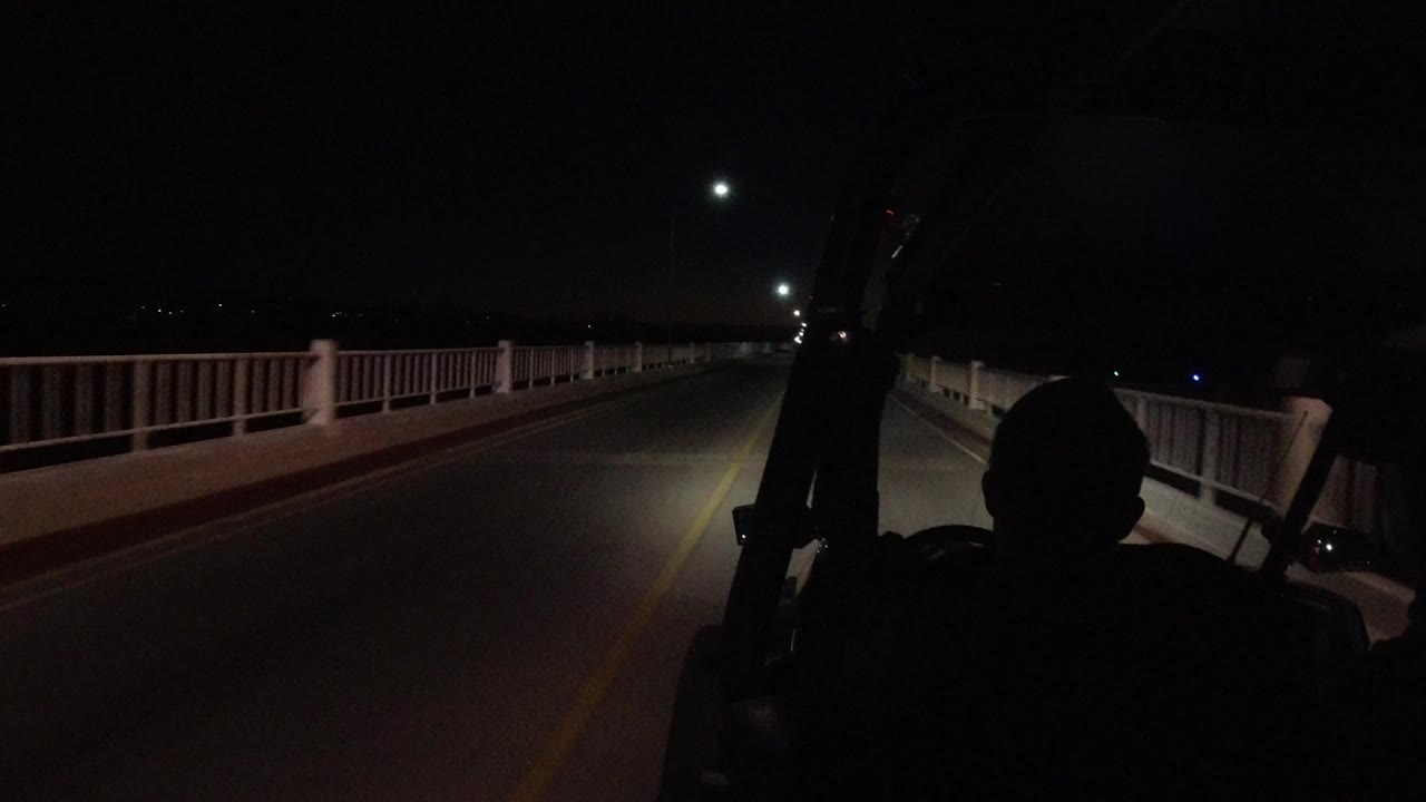 Dim streetlights and railings frame a night road from inside a vehicle.