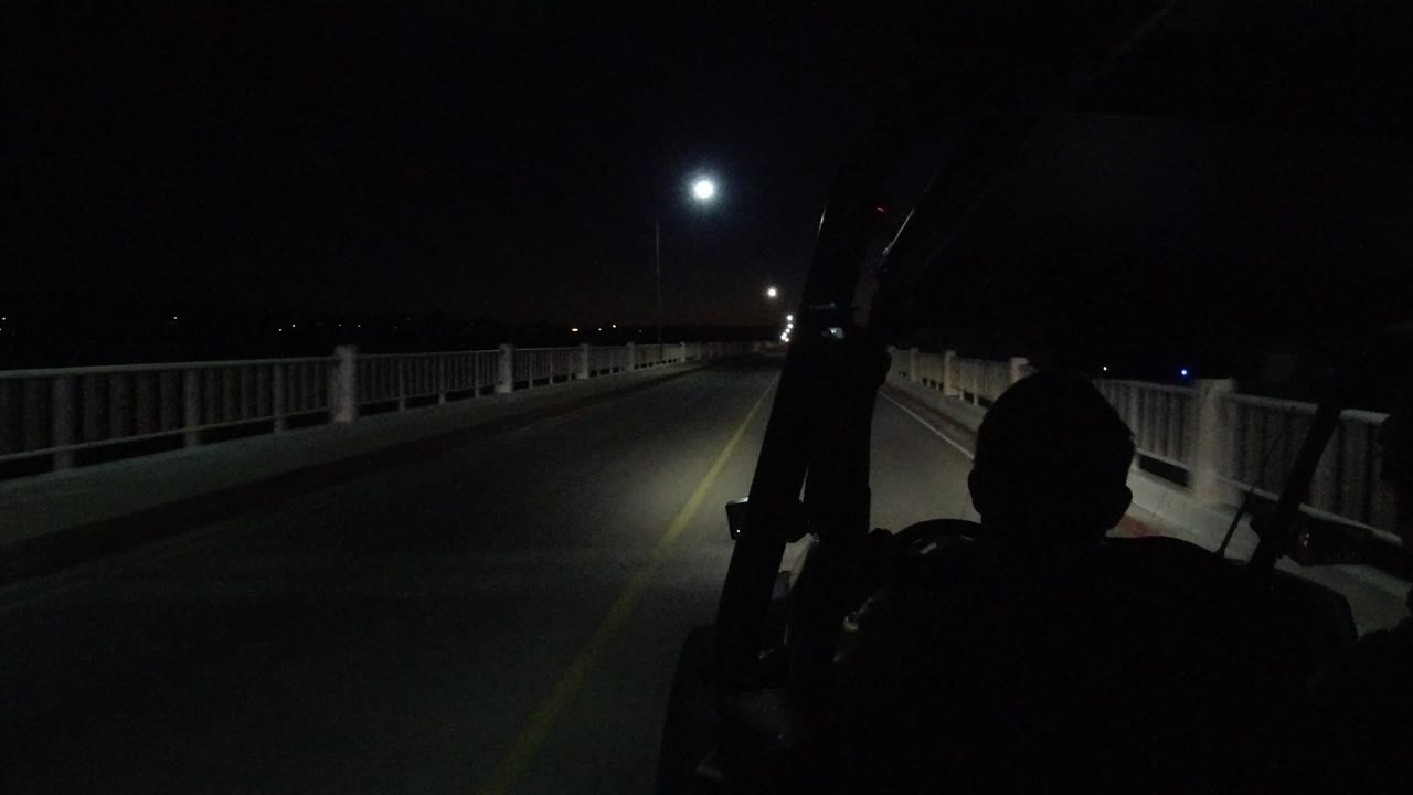 Opening view from a Jeep on a Los Cabos bridge at night, with a silhouetted person in the foreground, road railings, yellow lane markings, and a bright light ahead.