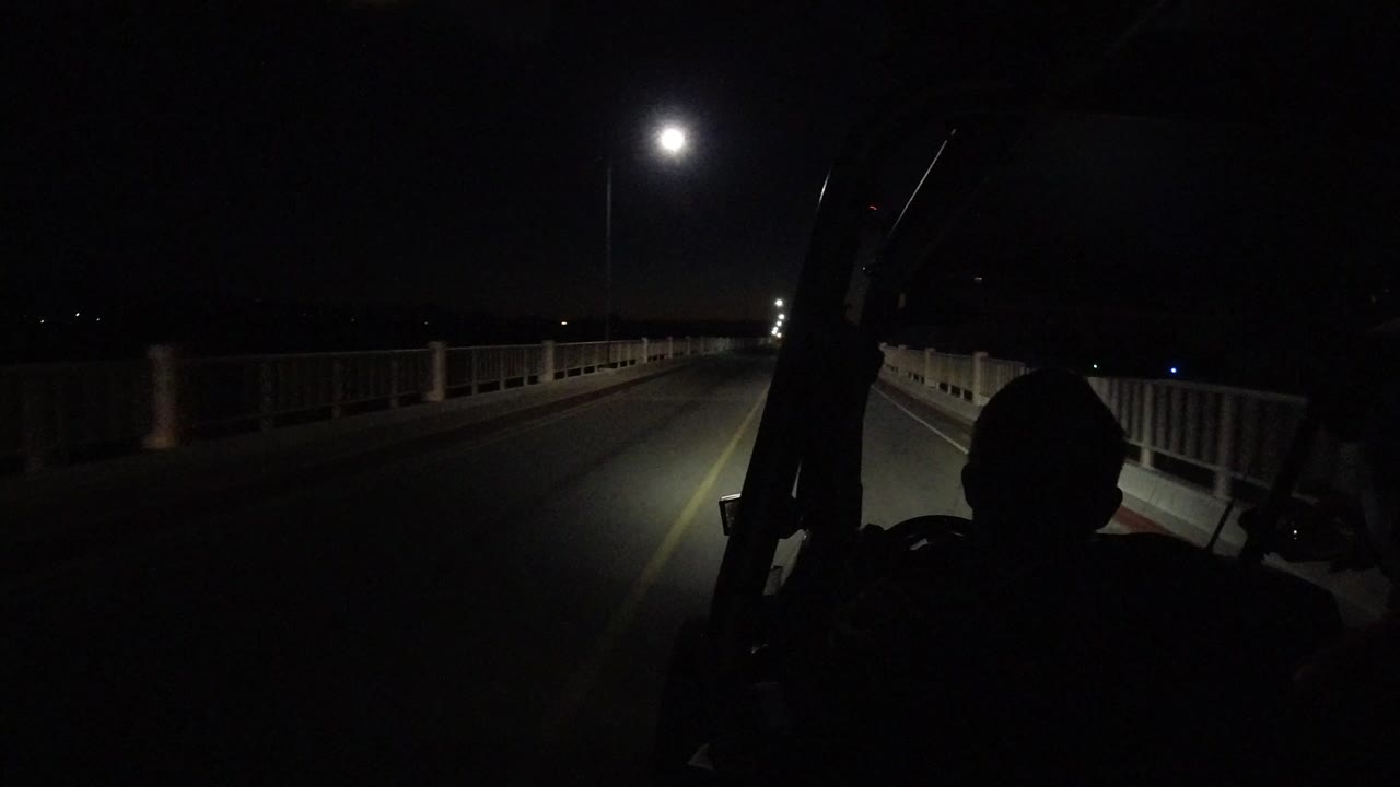 Start of a Los Cabos night bridge drive with railings on both sides, a yellow center line, headlight spill on the road, and a dark foreground occupant silhouette.