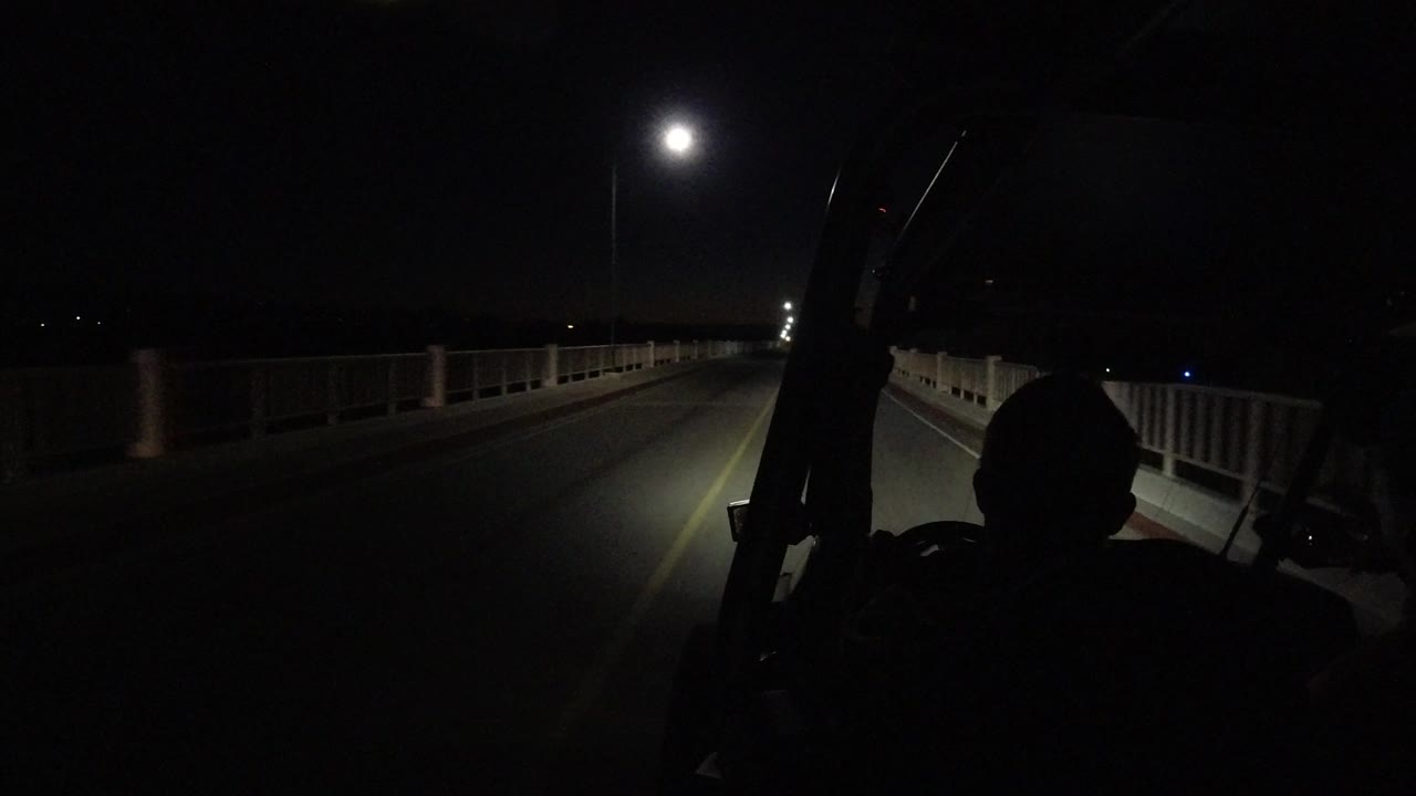 A Los Cabos bridge roadway continues into darkness, with headlight illumination along the right lane, side railings, distant lights, and the occupant silhouette low in frame.