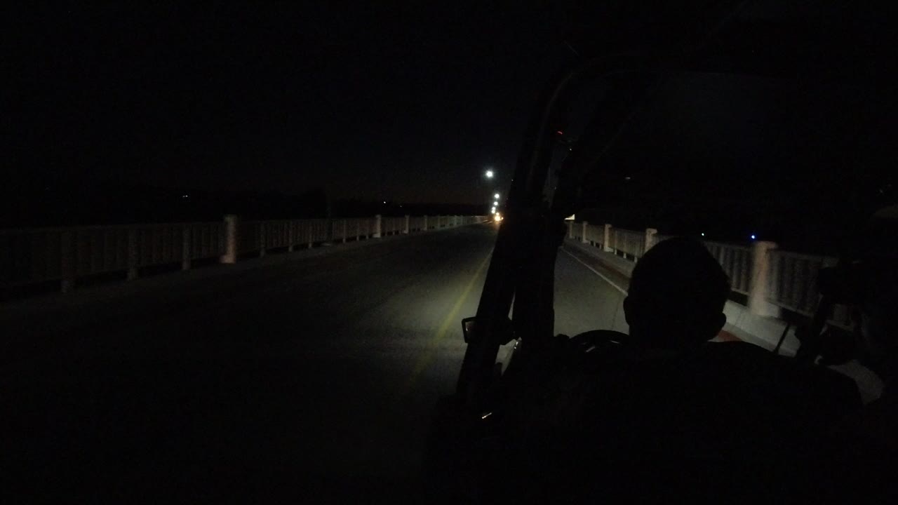 A dark Los Cabos road at night is seen from behind vehicle framing, with bridge-like railings, distant street lamps, and a silhouetted rider in the foreground.