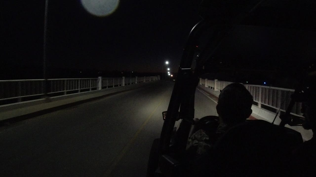 One second into the Los Cabos clip, the roadway remains centered under headlight spill, with railings on both sides and a silhouetted passenger shape at the lower right.