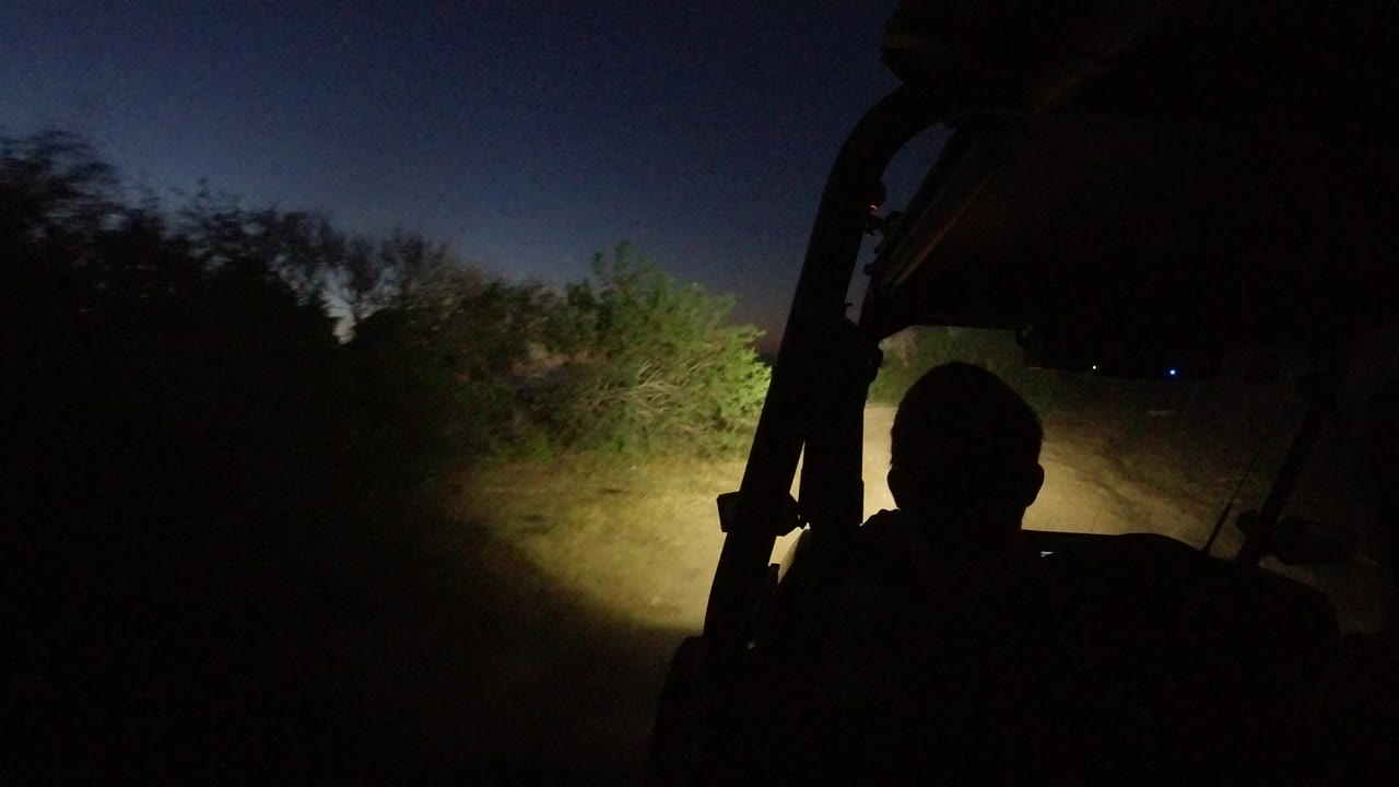 Dark vehicle interior facing a headlight-lit dirt road at dusk.