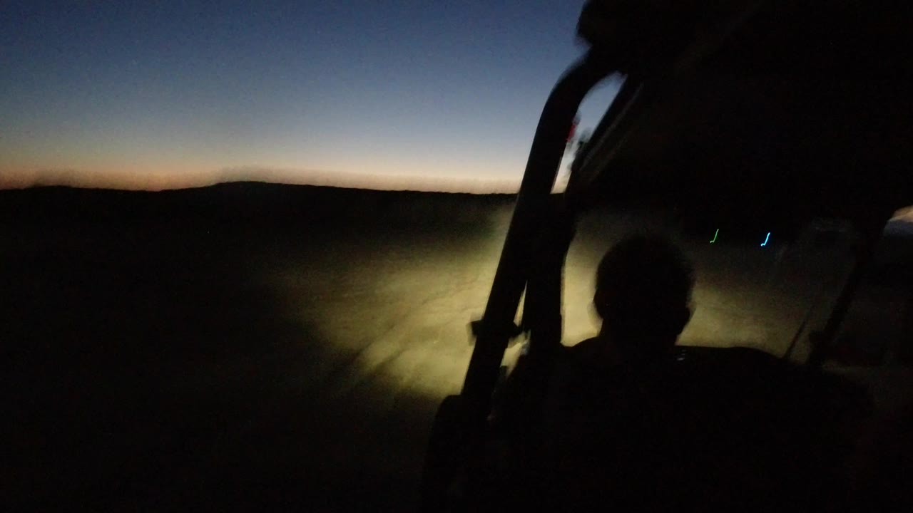 In Los Cabos, Mexico, a silhouetted person sits in an open vehicle as headlights illuminate a pale dirt track below a dark hill line and fading blue-orange sky.
