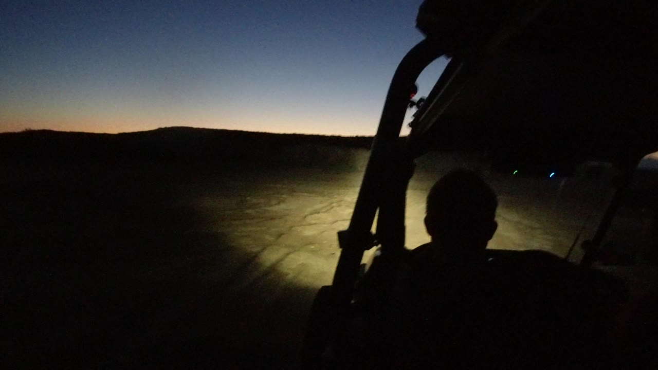 In Los Cabos, Mexico, the passenger silhouette stays centered while the vehicle lights spread across the uneven road surface and the horizon remains a thin warm band.