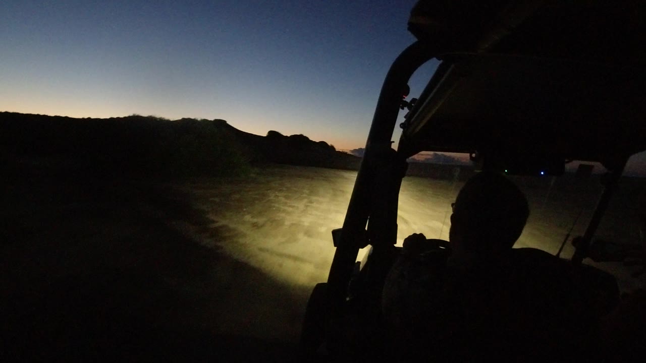 Los Cabos night road view from a vehicle, with the headlight beam spreading over the dirt surface and a dark occupant silhouette under the roof frame.