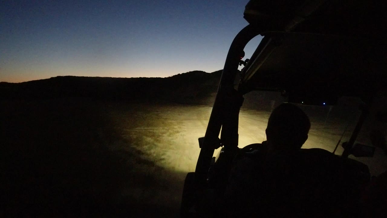 Opening frame from Los Cabos, Mexico, showing a dark vehicle interior silhouette, headlights on uneven rocky ground, low hills, and no recognizable faces.