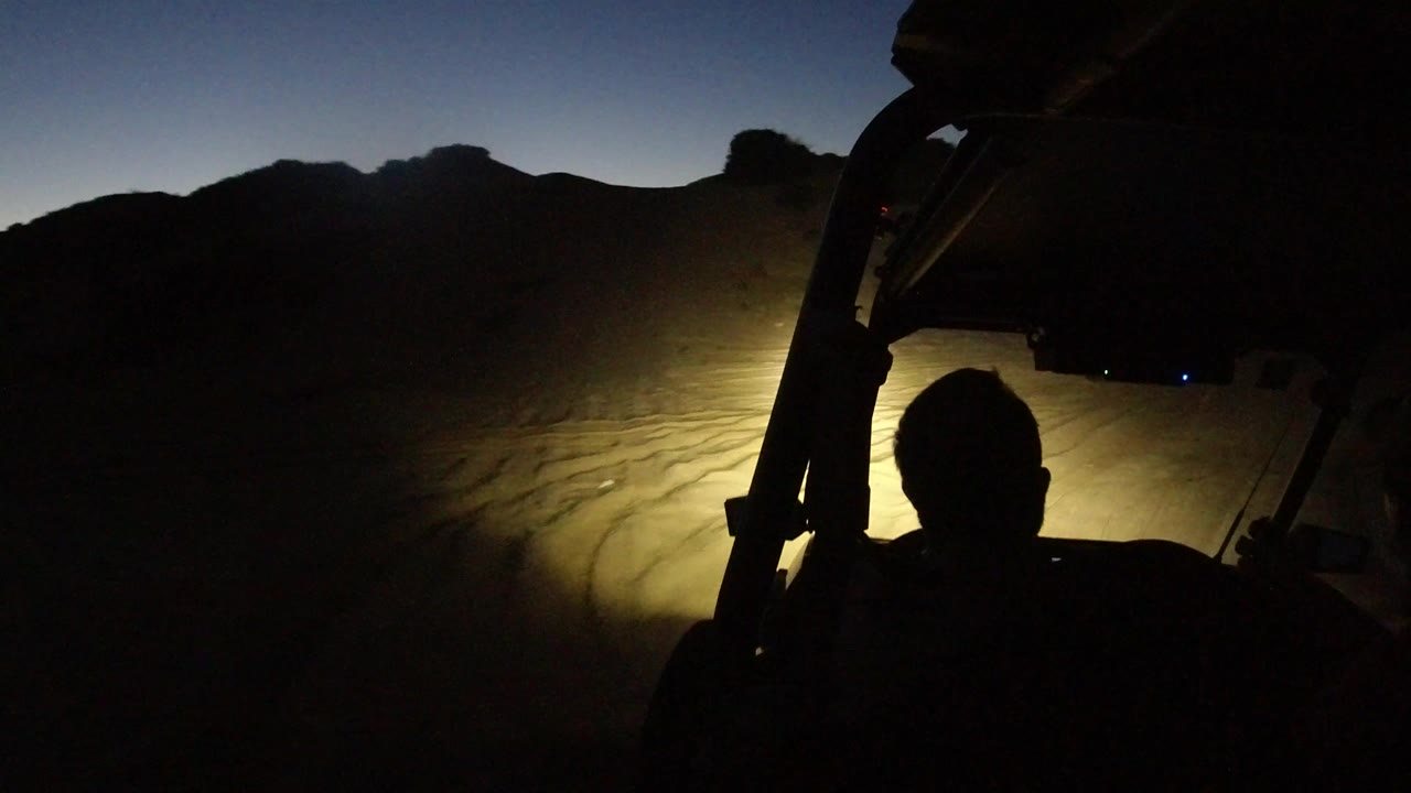 Dark beach track lit by vehicle headlights with a silhouetted cabin frame.