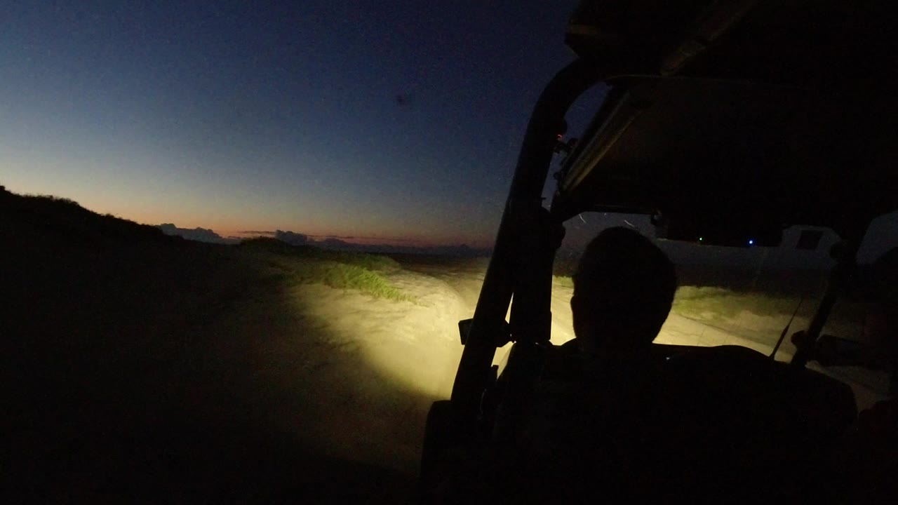 Interior Wrangler view in Los Cabos, Mexico, with a silhouetted person in front, headlights lighting a dirt track, and a fading blue-orange horizon.