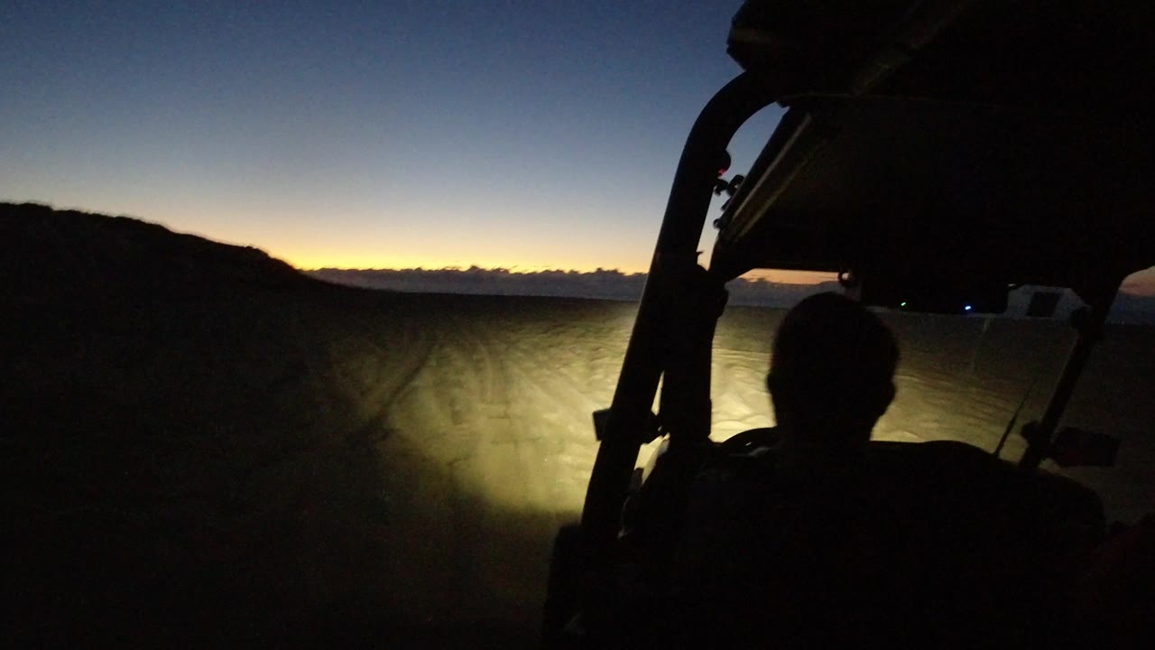 The driver remains in silhouette while the vehicle points toward sandy ruts, a low dark hillside, and an orange strip of sunset in Los Cabos, Mexico.