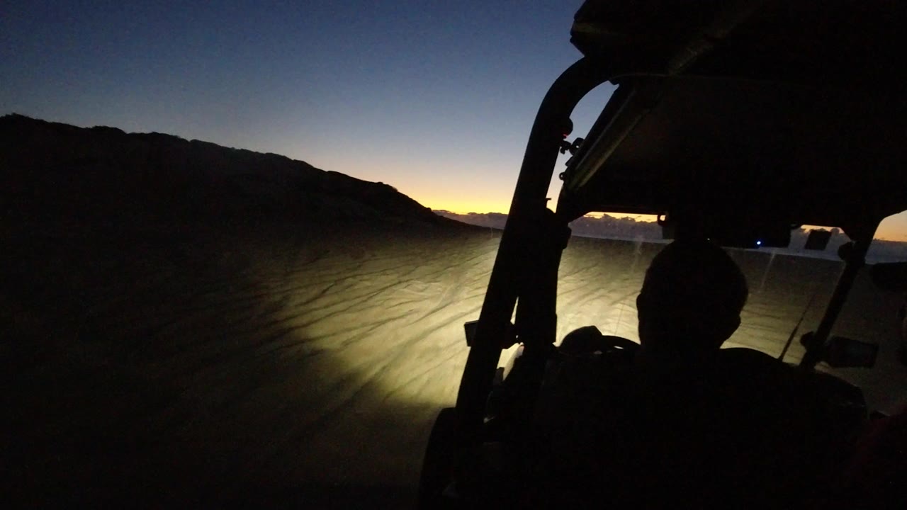 Inside-vehicle view in Los Cabos, Mexico, with a dark driver silhouette, roll bar, headlights on dusty ground, low hills, and no readable face.