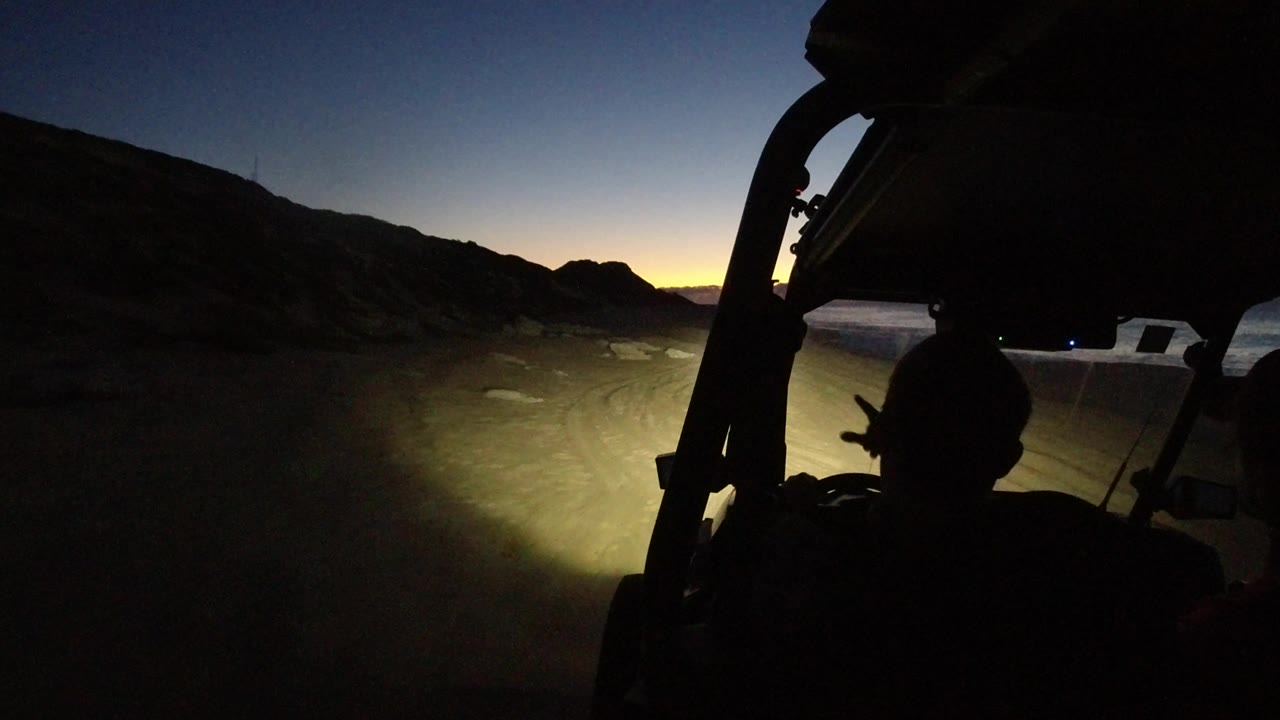 A silhouetted occupant sits below the vehicle roof frame as headlights reveal sandy ground, scattered rocks, dark hills, and a thin band of sunset color in Los Cabos.