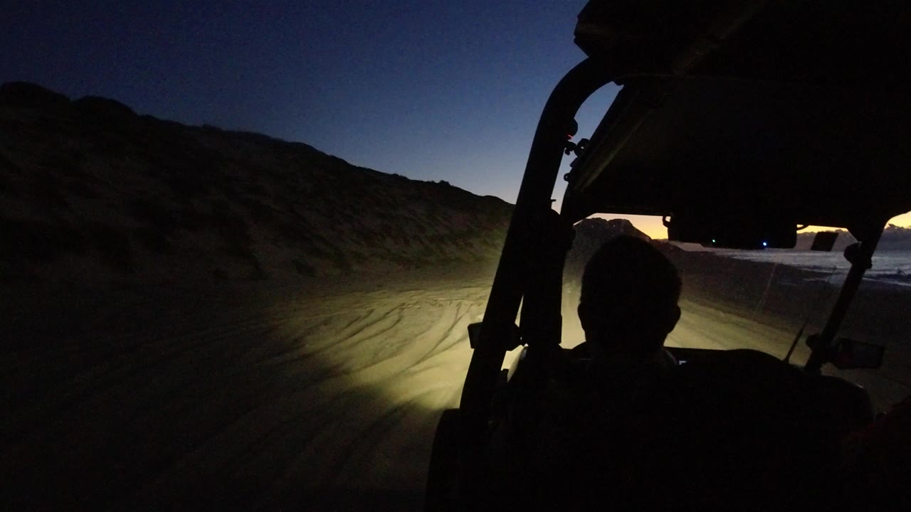 A silhouetted vehicle cabin looks forward in Los Cabos as headlights spread across the rutted road and a faint orange band sits low on the horizon.