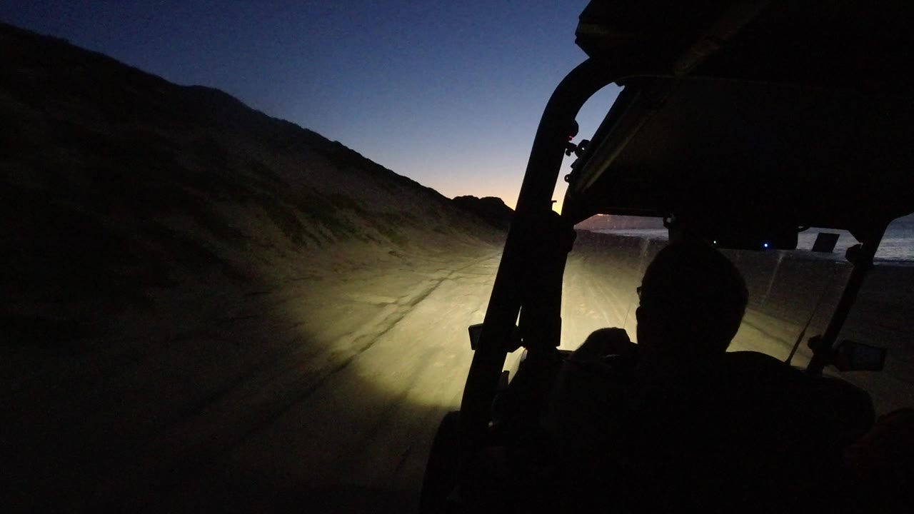 At 0:02 in Los Cabos, Mexico, the foreground is mostly black vehicle structure and driver silhouette, with headlights centered on a narrow dirt track beside a hillside.