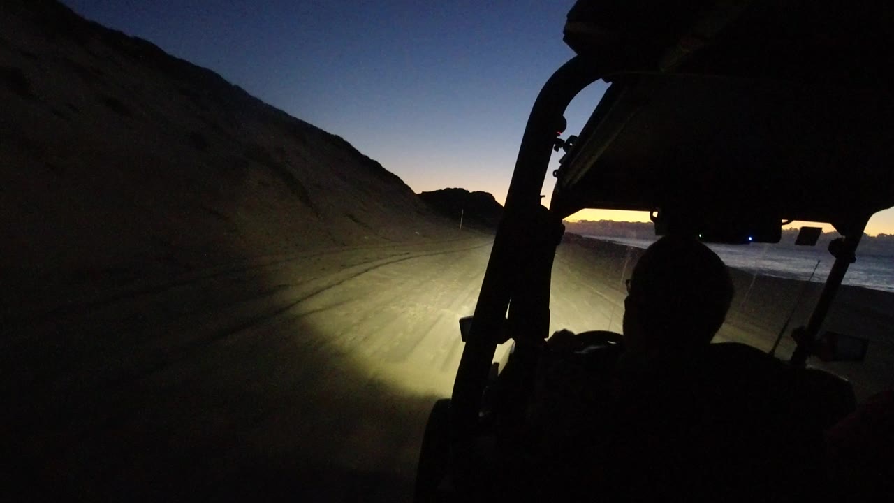 Opening frame from Los Cabos, Mexico showing a dark open-vehicle interior, a silhouetted occupant, and headlights lighting a sandy path near the coast at dusk.