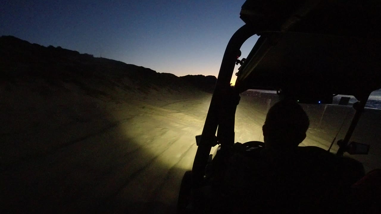 Dark vehicle interior facing a headlit dirt road at dawn.