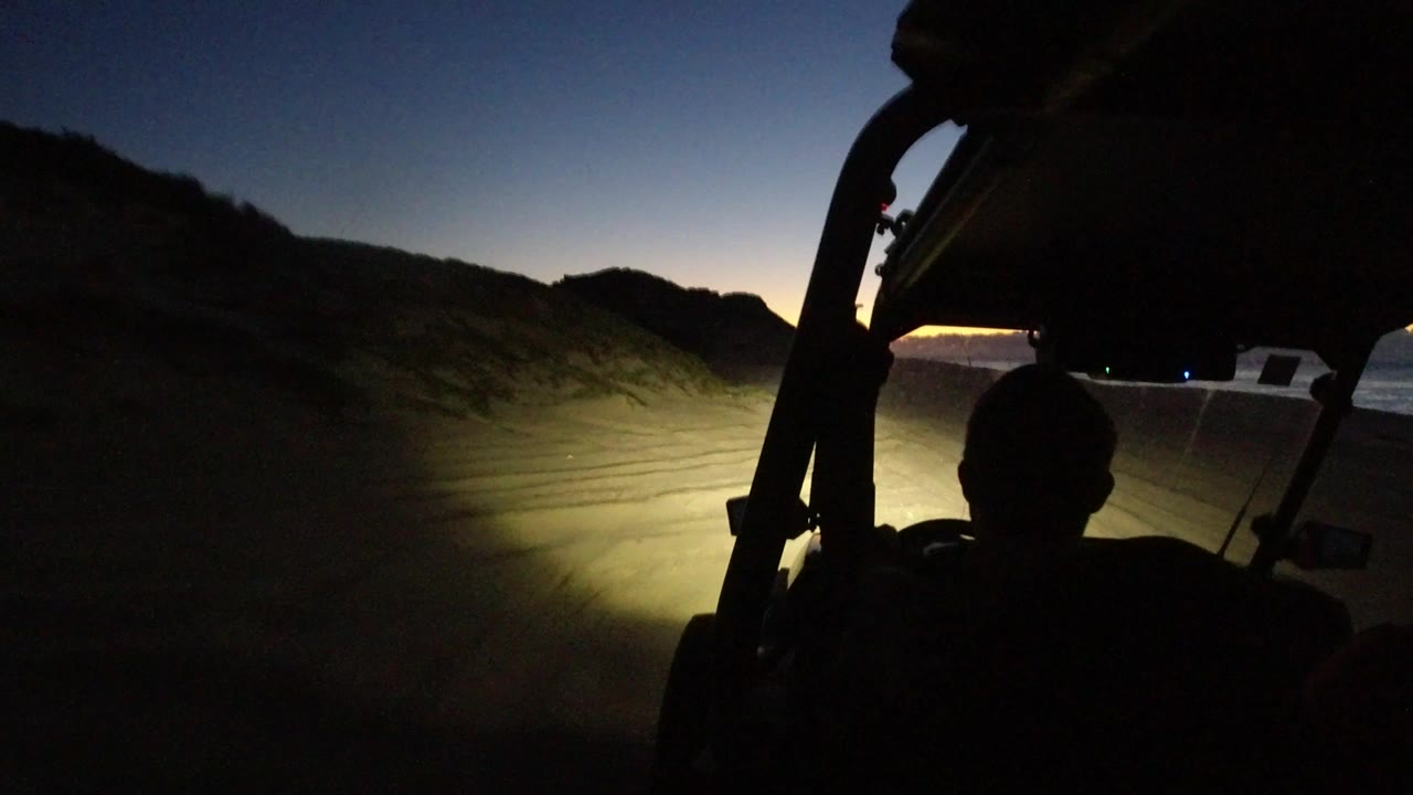 Silhouetted off-road vehicle interior facing headlights on desert sand at dusk.