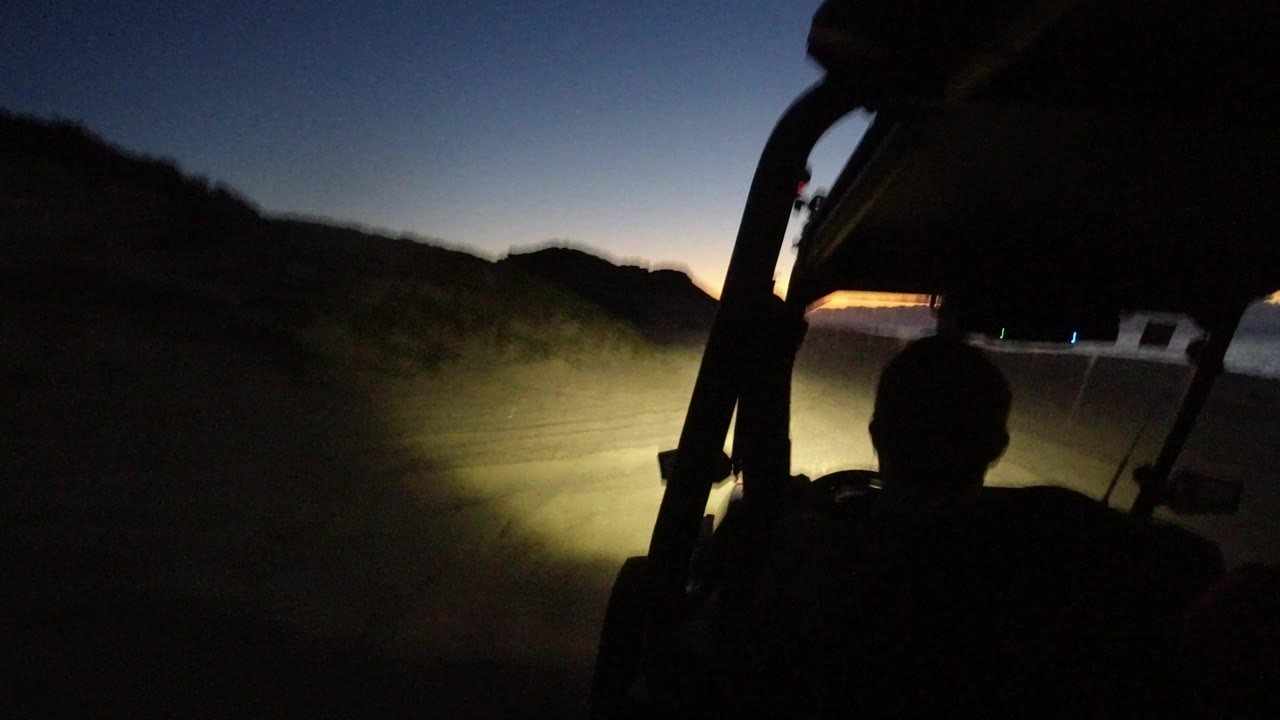 At 0:01, the forward view from the vehicle shows the driver's dark silhouette, a windshield edge, bright headlights on sand, and low hills against the remaining horizon glow in Los Cabos.