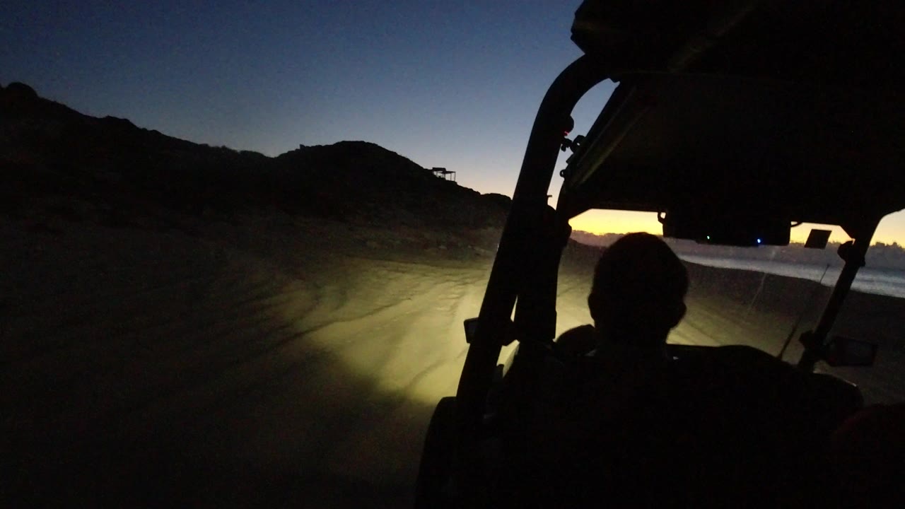 Silhouetted rider inside a vehicle at dawn with headlights on sandy ground